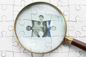 A magnifying glass hovering over a dollar bill that’s hidden behind several puzzle pieces
